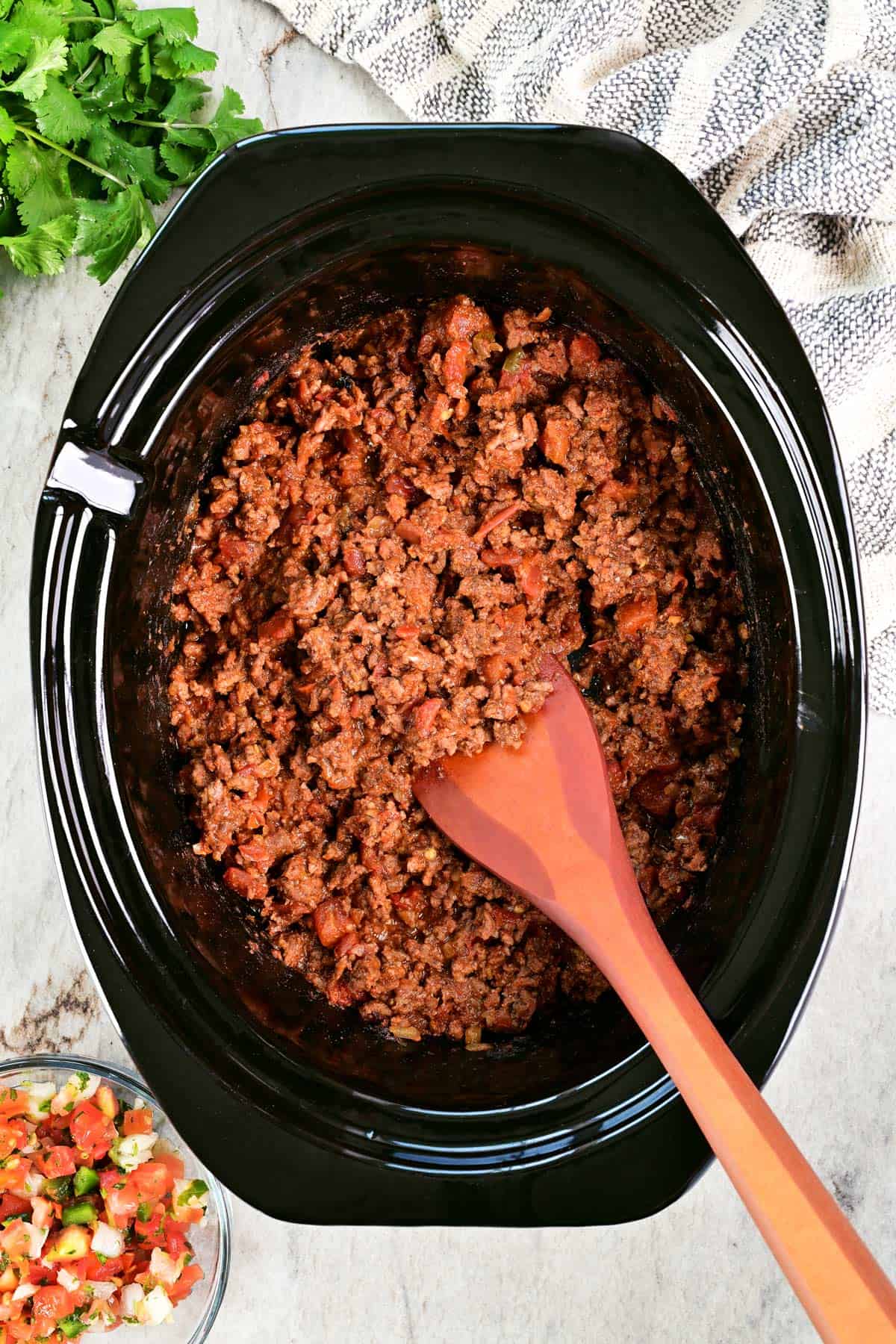 Slow cooker tacos with a wooden spoon and toppings on the side.