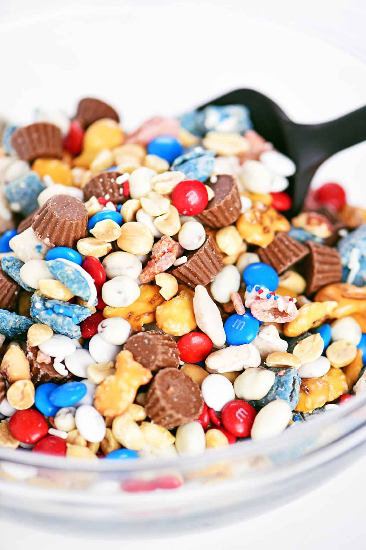 Red white and blue trail mix in a glass bowl with a black silicone spatula.