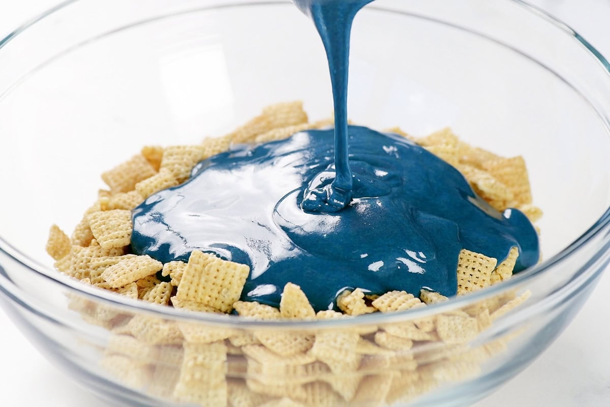 Pouring blue coating over Rice Chex in a glass bowl.