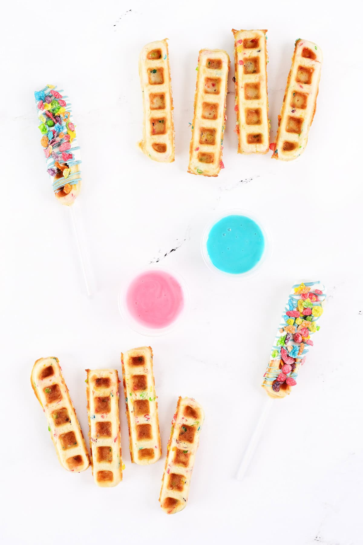 Fruity pebbles waffle sticks and pops with pink and aqua frosting in cups.