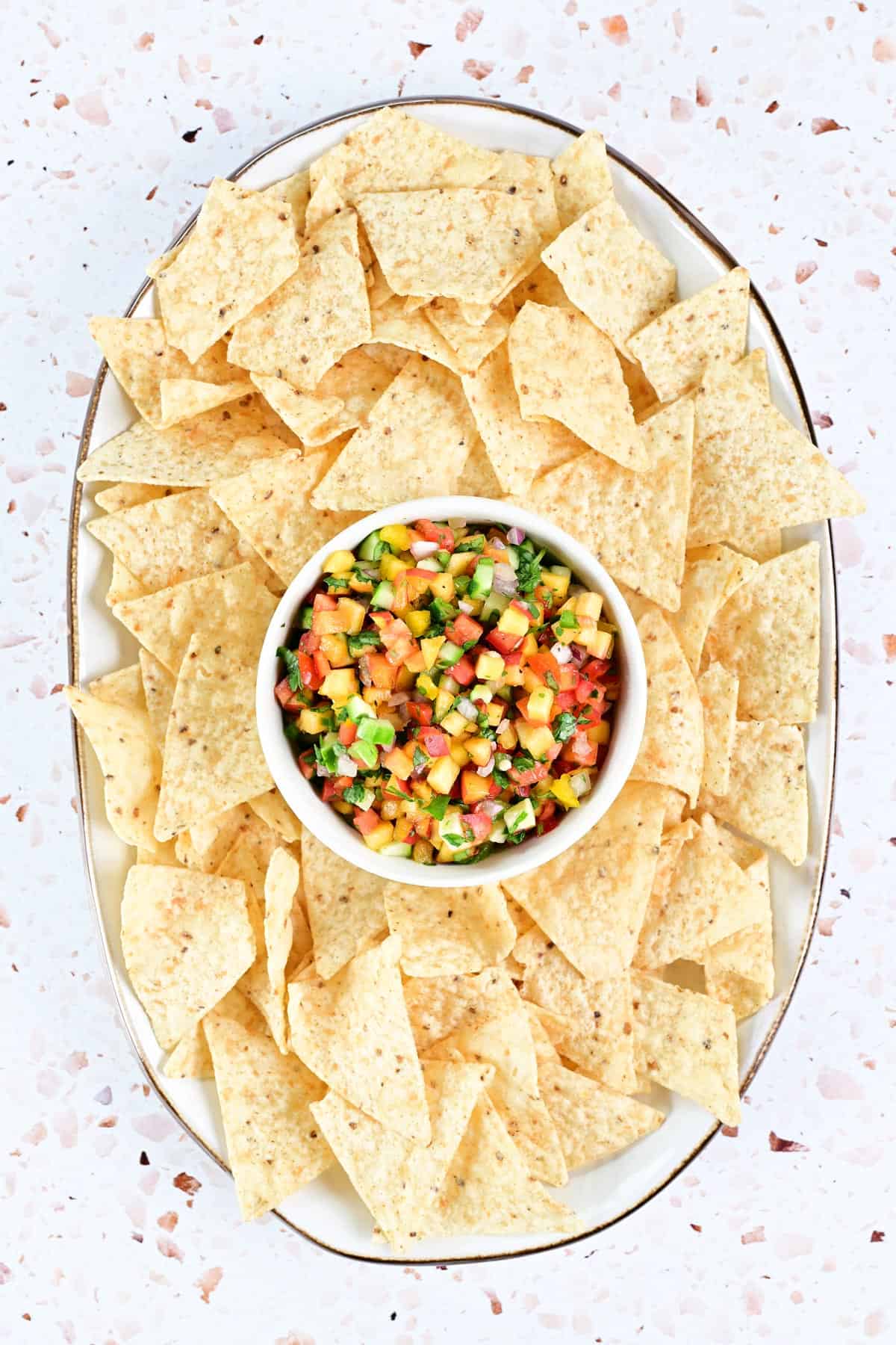 Peach salsa in a white bowl on an oval tray surrounded by tortilla chips.