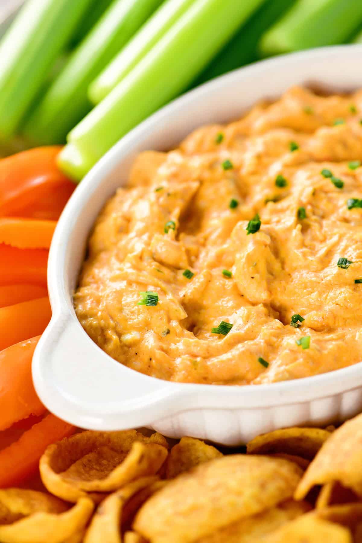 Slow cooker buffalo chicken dip in a white dish surrounded by celery, peppers, and chips.